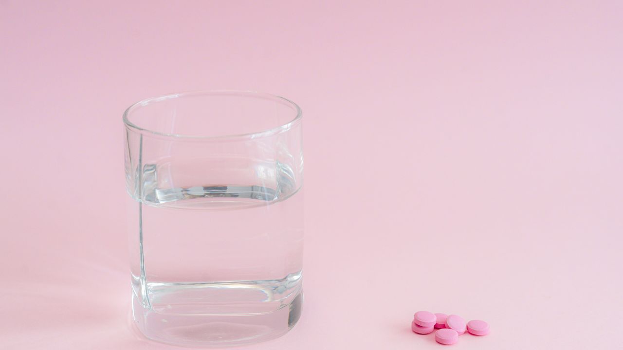 Glass of water next to pink tablets on a pink background